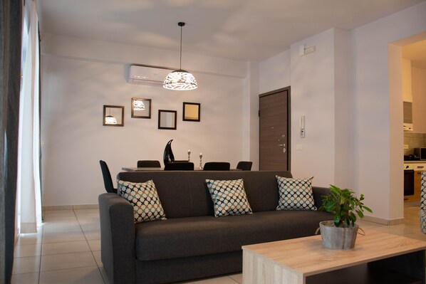 Een moderne woonkamer met een bank, salontafel en decoratieve kussens. Er is een eethoek met stoelen en een tafel, en een keuken op de achtergrond.
