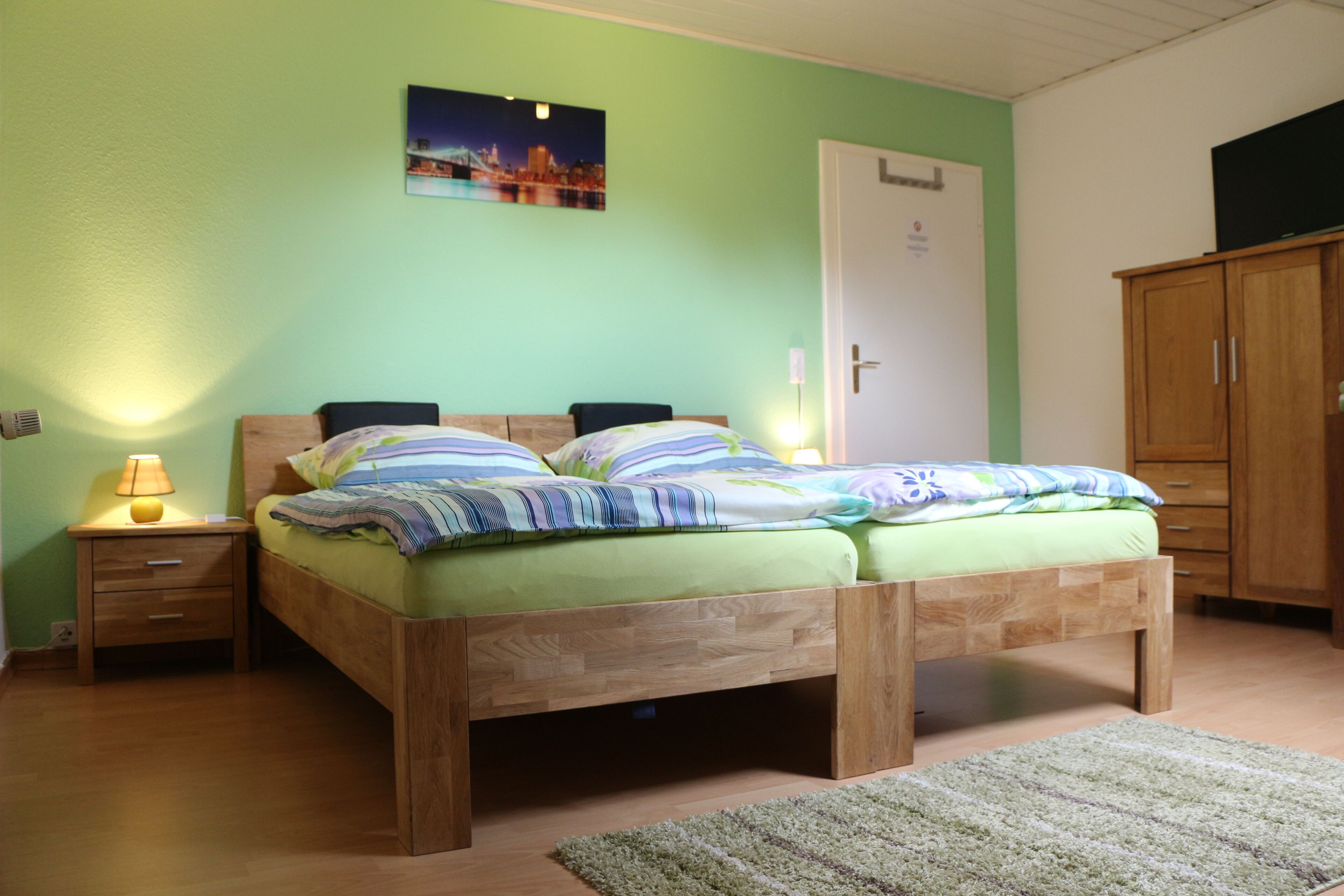 A bedroom with two wooden beds, a nightstand with a lamp, a green wall, a door, and a wardrobe.