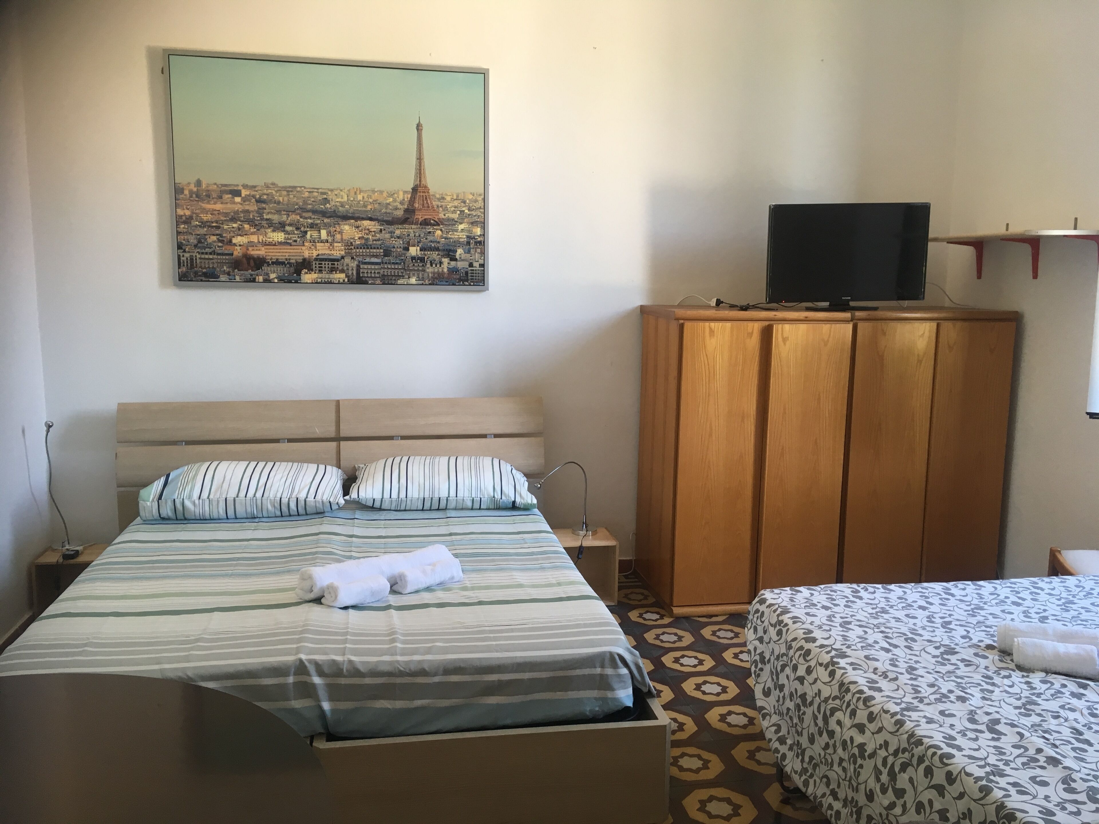 A hotel room with a bed, a TV on a wooden cabinet, and a painting of the Eiffel Tower on the wall.