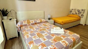 A bedroom with a large bed, a smaller bed, a nightstand, a lamp, a potted plant, and a painting on the wall.