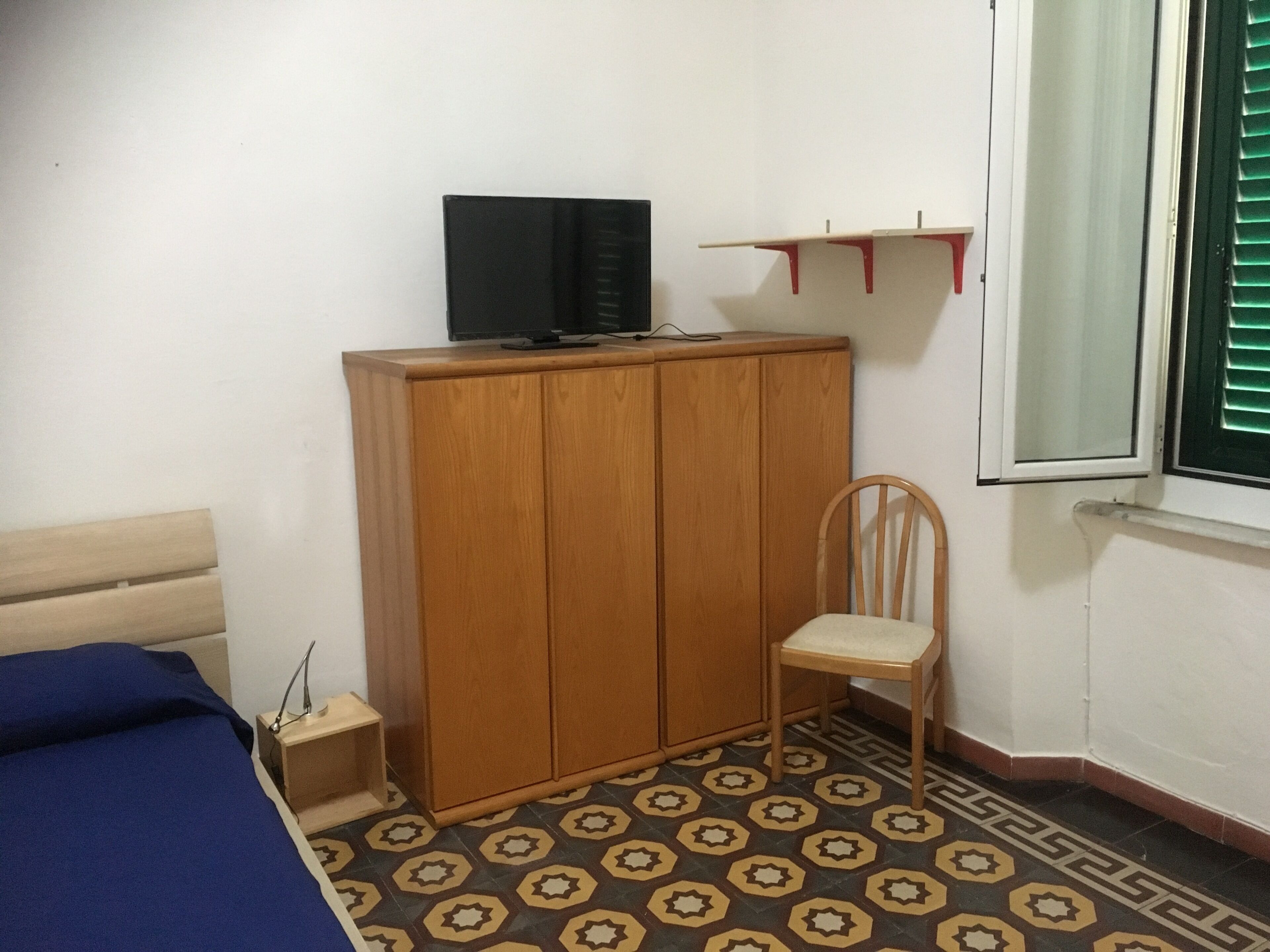 Une chambre avec une armoire en bois, un lit simple recouvert d’une couverture bleue, une chaise et un téléviseur posé sur le dessus de l’armoire.