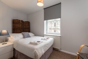 Un dormitorio ordenado con cama, lámpara de noche y ventana con vistas a edificios.