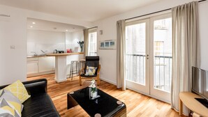 A modern living room with a black sofa, a coffee table, and a TV stand. There is a kitchen area with a bar stool and a chair. The room has large windows with curtains and a sliding glass door leading to a balcony. - Silver Lining - Grassmarket Apartments (Edinburgh)
