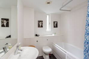 Un baño moderno con lavamanos, inodoro y bañera blancos. Hay cuadros enmarcados en la pared, una ventana sobre el inodoro y una cortina de ducha con un estampado azul.