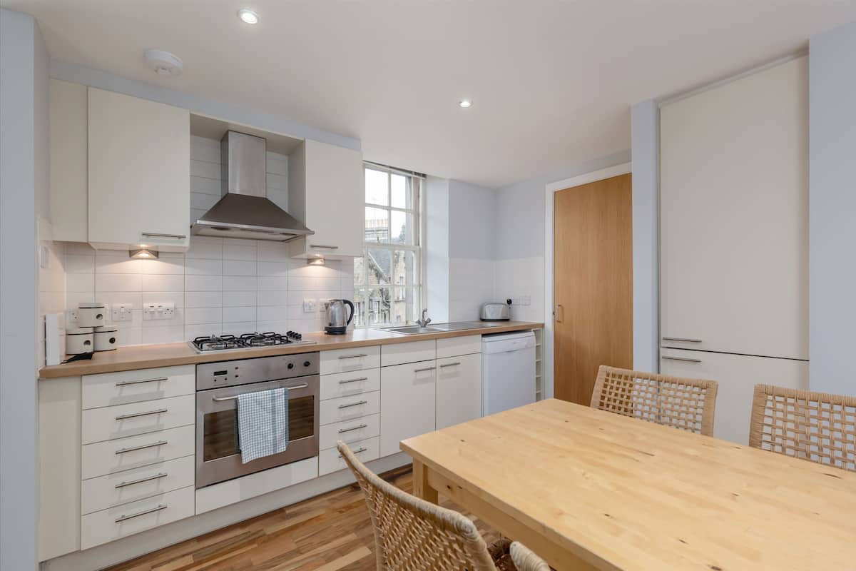 A modern kitchen with white cabinets, a wooden countertop, a built-in oven, and a dining area with a wooden table and wicker chairs.