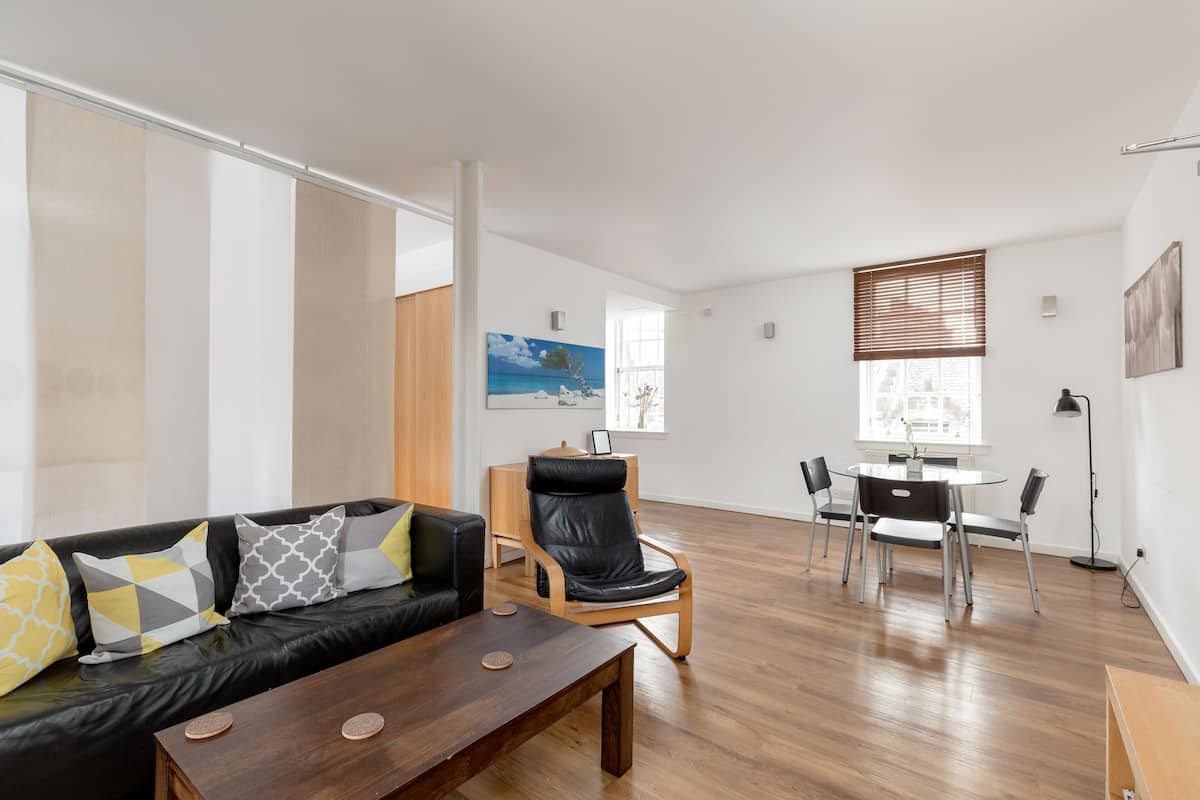 A modern living room with a black leather sofa, a wooden coffee table, and a dining area with a round table and chairs.