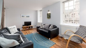 A modern living room with a black leather sofa, a wooden coffee table, a TV, and a large window with a city view. - Silver Lining - Grassmarket Apartments (Edinburgh)