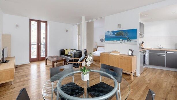 A modern living room with a glass dining table, a sofa, and a kitchen area with grey cabinets and a wooden countertop. - Silver Lining - Grassmarket Apartments (Edinburgh)