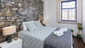 A bedroom with a stone wall, a bed with patterned bedding, a window with shutters, and towels on the bed.