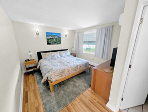 A bedroom with a bed, bedside tables, a nightstand, a TV, a window with curtains, and a painting on the wall.