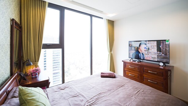 A hotel room with a large bed, a TV on a wooden dresser, a window with curtains, and a view of buildings outside. - Luxury Apartment Vinhomes Metropolis 02 BR#5**Sky View# (Hà Nội)