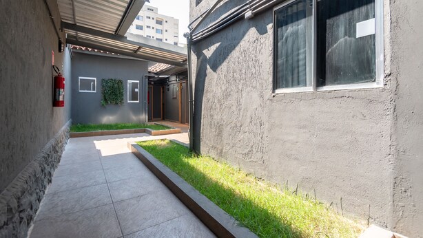 A narrow pathway with a fire extinguisher on the left, a small patch of grass, and a building with a textured wall and windows. - OYO Hotel Massimo Brooklin, Sao Paulo (São Paulo)