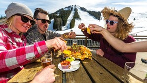A group of people dining outdoors at a wooden table with a view of a snowy mountain. - Perfect for larger groups, fantastic view to Red Mountain (Rossland)