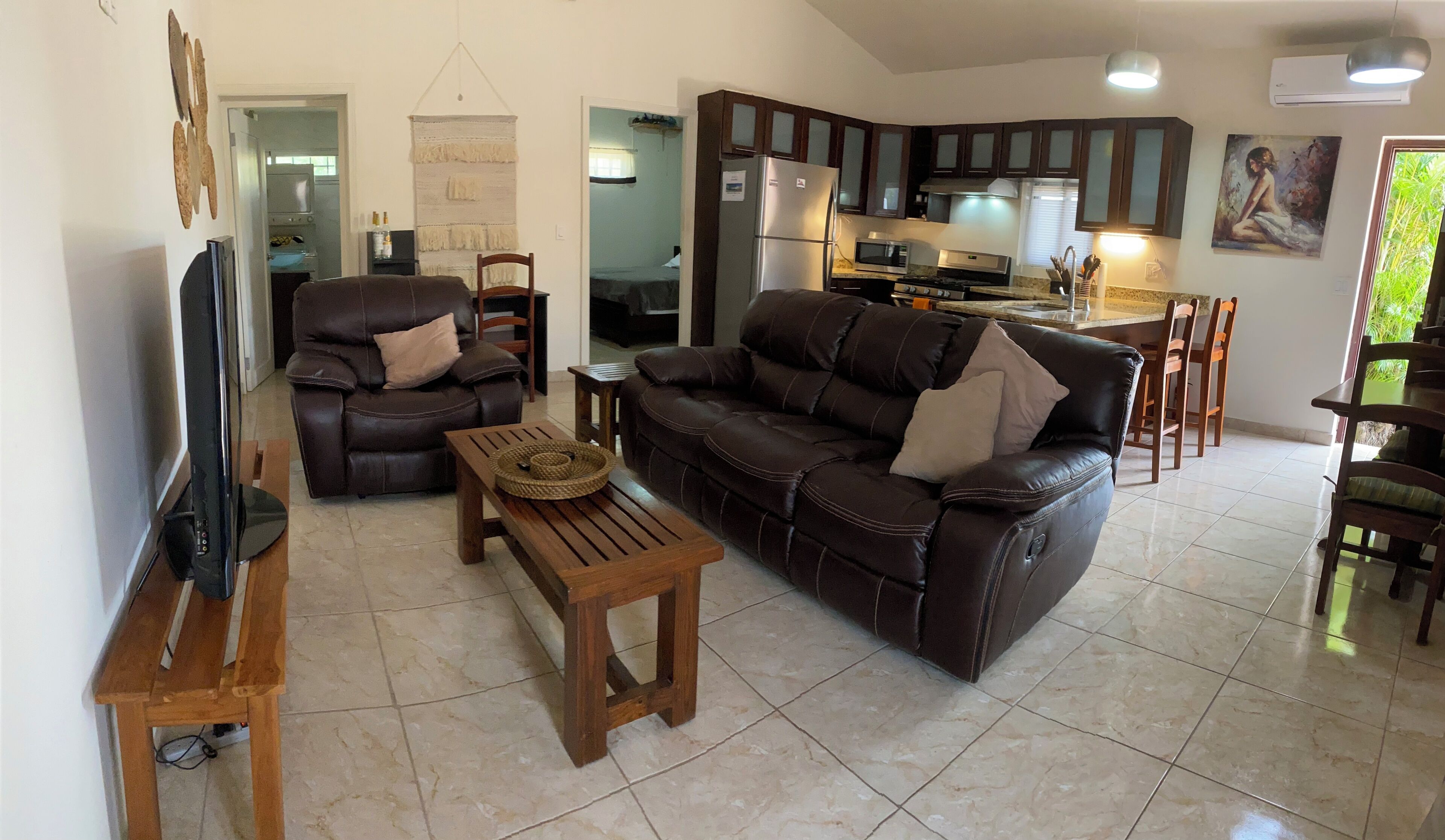 A living room with a leather sofa, a wooden coffee table, a TV, and a kitchen in the background.
