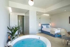 A modern hotel room with a large bathtub, a bed with a blue headboard, and a small table with a chair.