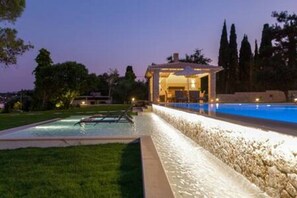 A swimming pool with a stone wall, a covered seating area, and a view of trees and a building in the background.
