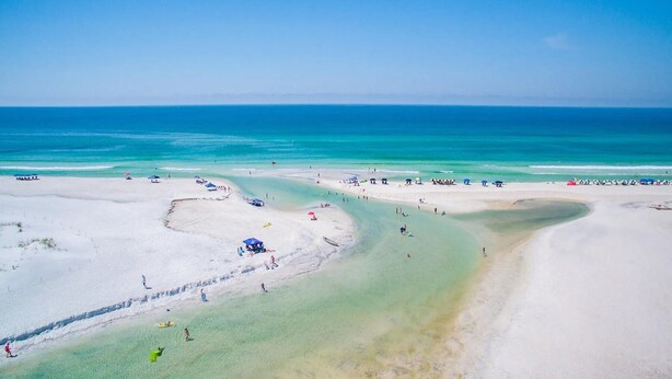 Ein Strand mit klarem türkisblauem Wasser, Sandküste und mehreren Sonnenschirmen und Liegestühlen.