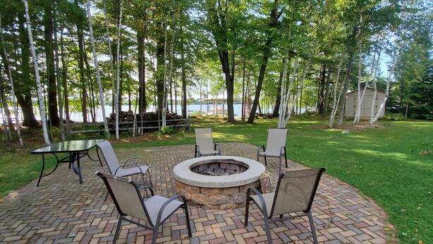A circular fire pit with four chairs around it, set on a brick patio with a view of a lake and trees in the background. - Sunset Cove Eagle River- Waterfront Retreat on Catfish Lake (Eagle River)
