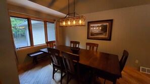 Un coin repas avec une table et des chaises en bois, un luminaire suspendu, une fenêtre avec des stores et un tableau encadré au mur.