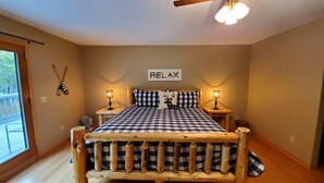 A bedroom with a checkered bedspread, two bedside tables with lamps, a ceiling fan, and a 'RELAX' sign on the wall. - Sunset Cove Eagle River- Waterfront Retreat on Catfish Lake (Eagle River)