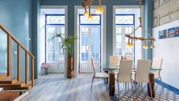 Une pièce spacieuse avec un escalier en bois, un coin repas comprenant des chaises et une table en verre, et une grande fenêtre dotée de volets bleus.
