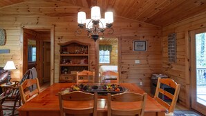 A wooden cabin interior with a dining table, chairs, a chandelier, a bookshelf, and a sign on the wall.