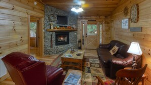 A cozy cabin interior with a stone fireplace, wooden walls, a leather sofa, and a television mounted above the fireplace.