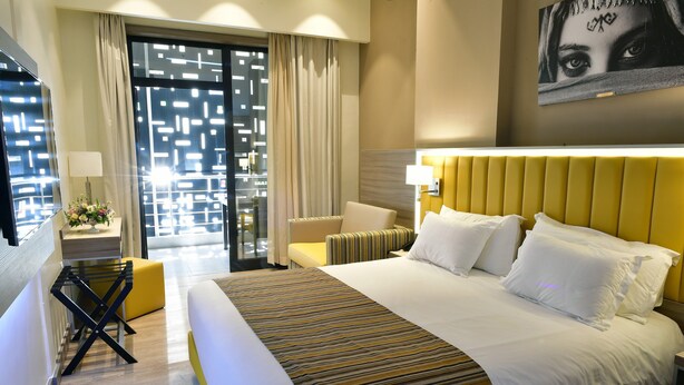 A hotel room with a large bed, a yellow headboard, a small table with a lamp, and a view through a glass door. - Naya (Algiers)