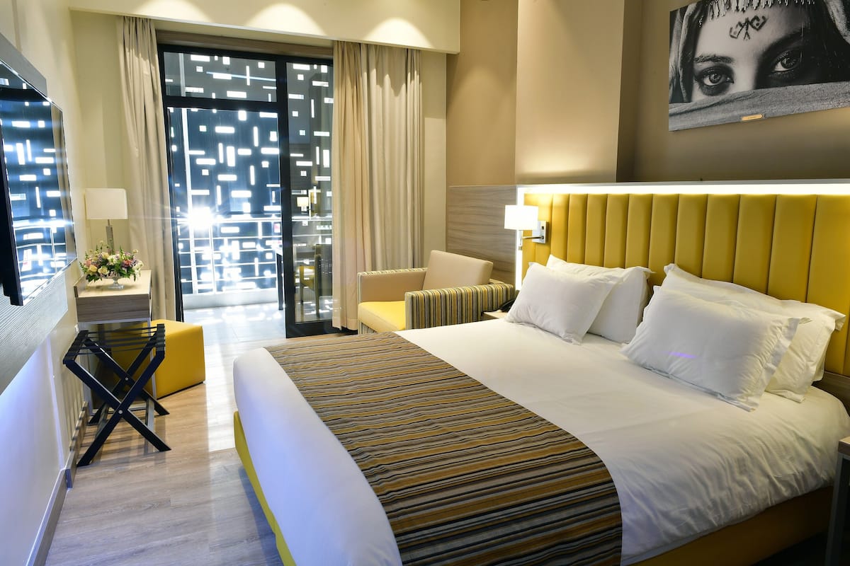 A hotel room with a large bed, a yellow headboard, a small table with a lamp, and a view through a glass door.