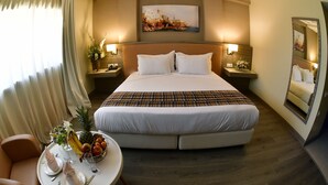 A hotel room with a large bed, bedside tables with lamps, a dining table with a fruit bowl, and a painting on the wall. - Naya (Algiers)