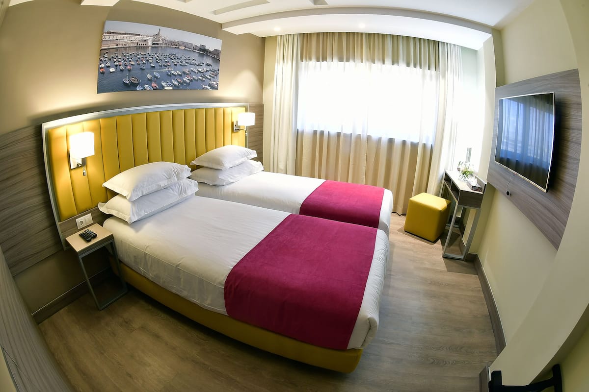 A hotel room with two beds, a TV, a nightstand, and a painting of a cityscape.