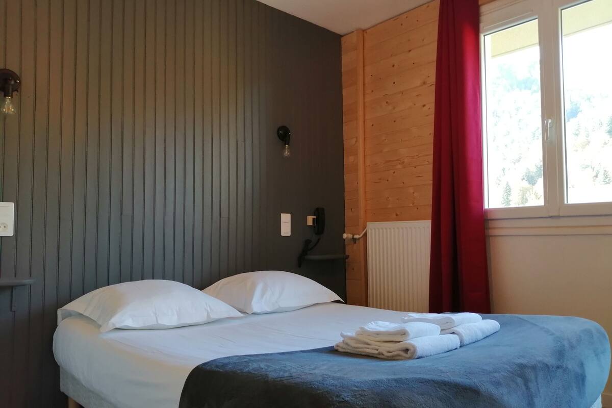 Une chambre à coucher avec un lit, une table de chevet, une lampe, une fenêtre avec des rideaux et un radiateur.