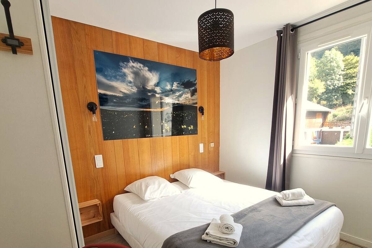 Une chambre à coucher avec une tête de lit en bois, un tableau représentant un ciel nuageux, un lit avec du linge de lit blanc et une couverture grise, une chaise rouge et une fenêtre avec des rideaux.