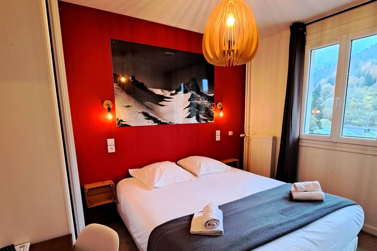 Une chambre d’hôtel avec un lit, un mur rouge, un tableau représentant une montagne et une fenêtre donnant sur un paysage.