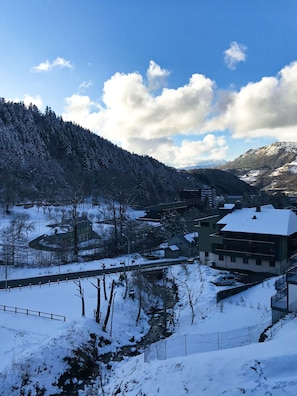 A snow-covered landscape with a river, buildings, and a forested mountain. - Hôtel Les Mouflons (Mont-Dore)