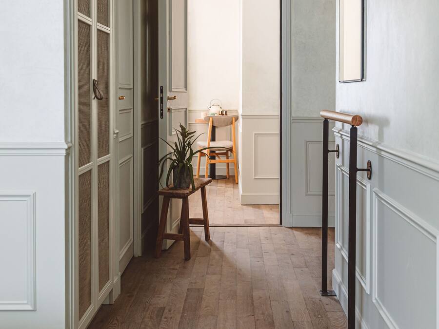 Un pasillo con piso de madera, paredes blancas y un espejo a la derecha. Se ve una puerta que da a otra habitación y una pequeña mesa con una planta.