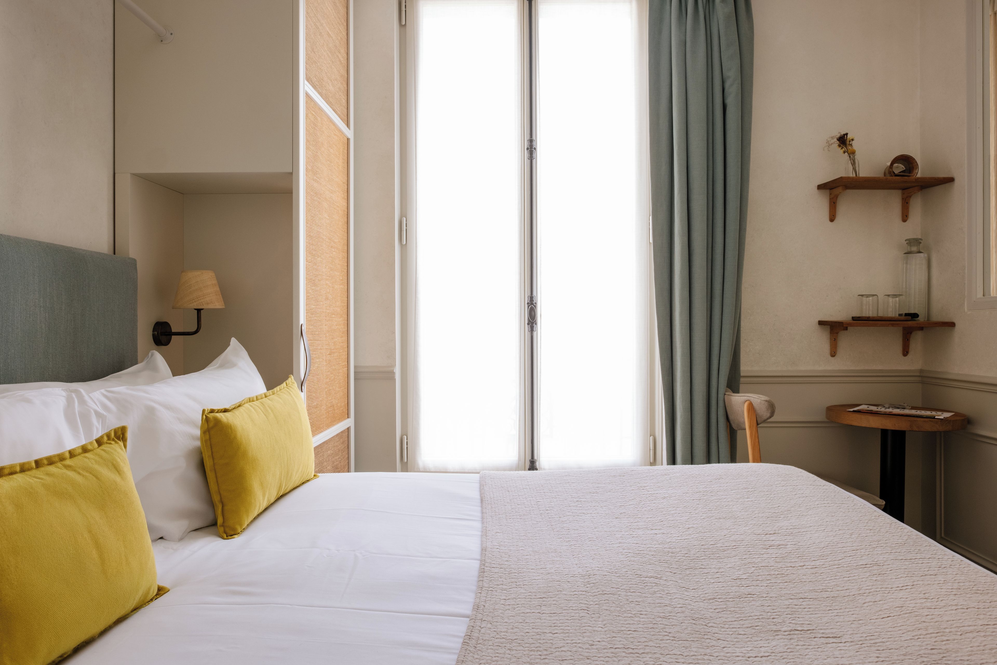 A hotel room with a bed, a nightstand, a lamp, a window with curtains, and a small table with a chair.