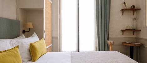 A hotel room with a bed, a nightstand, a lamp, a window with curtains, and a small table with a chair.