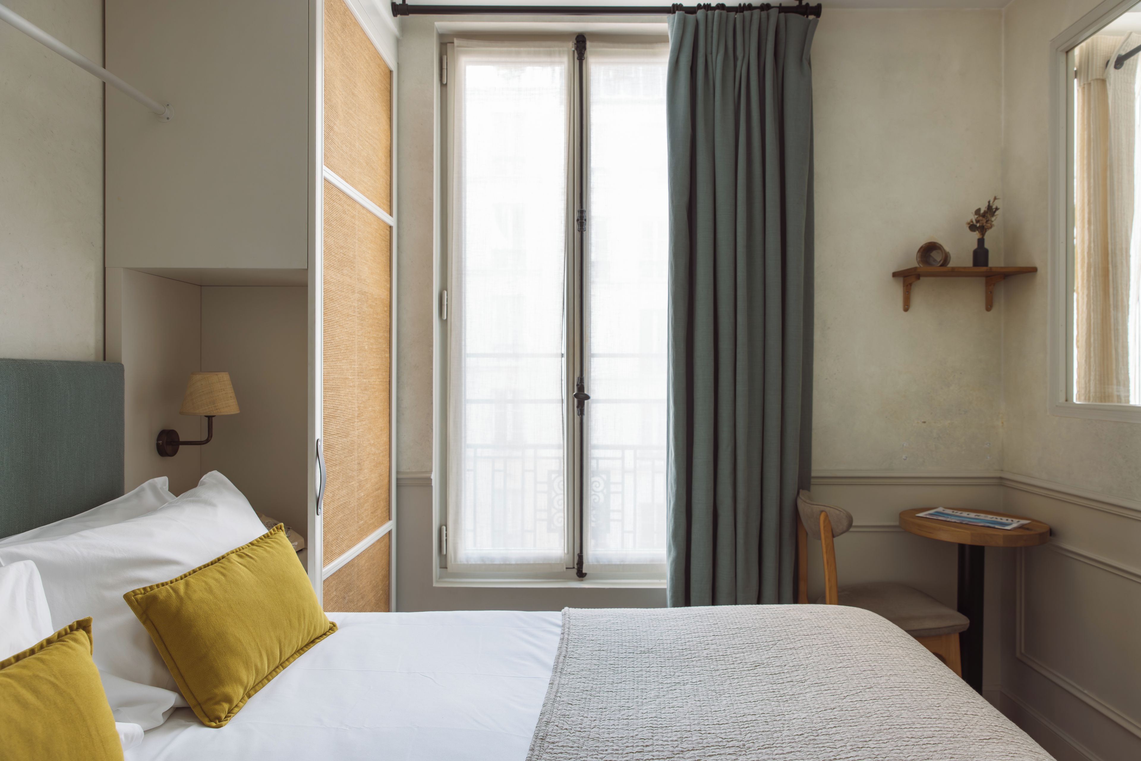 Una habitación de hotel con una cama, un ventanal con cortinas, una mesita y un estante en la pared.