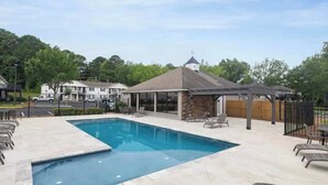 A swimming pool with lounge chairs, a covered seating area, and a building with a chimney in the background.