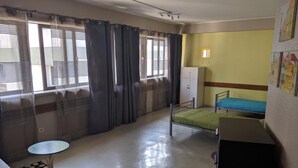 A small, well-lit room with a bed, a desk, and a chair. There is a window with curtains, a radiator, and a wall-mounted light fixture. - City Drops Hostel (Porto)