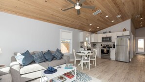 A modern living room with a sofa, coffee table, and a dining area with chairs. There is a kitchen with white cabinets, a microwave, and a refrigerator.