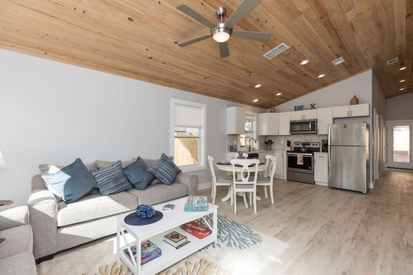 A modern living room with a sofa, coffee table, and a dining area with chairs. There is a kitchen with white cabinets, a microwave, and a refrigerator.