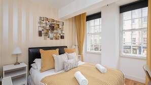 A hotel room with a bed, a nightstand, a wall art piece, a window with curtains, and a view of buildings outside. - Silver Lining - Luxury Jackson Apartment (Edinburgh)