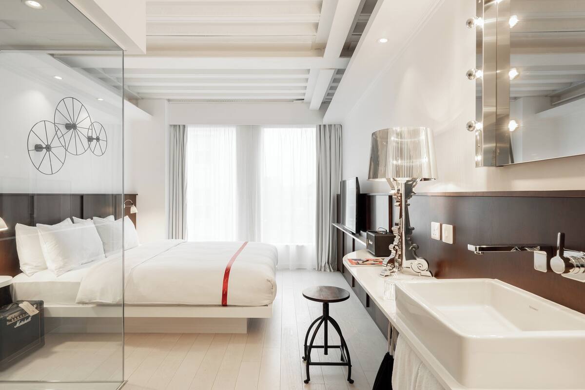 Ein modernes Hotelzimmer mit Bett, Nachttisch, Lampe, Spiegel und einem geräumigen Badezimmer mit Badewanne.