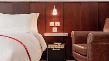 Ein Hotelzimmer mit Bett, einem kleinen Tisch mit Lampe, einem Ledersessel und einem Marshall Verstärker.