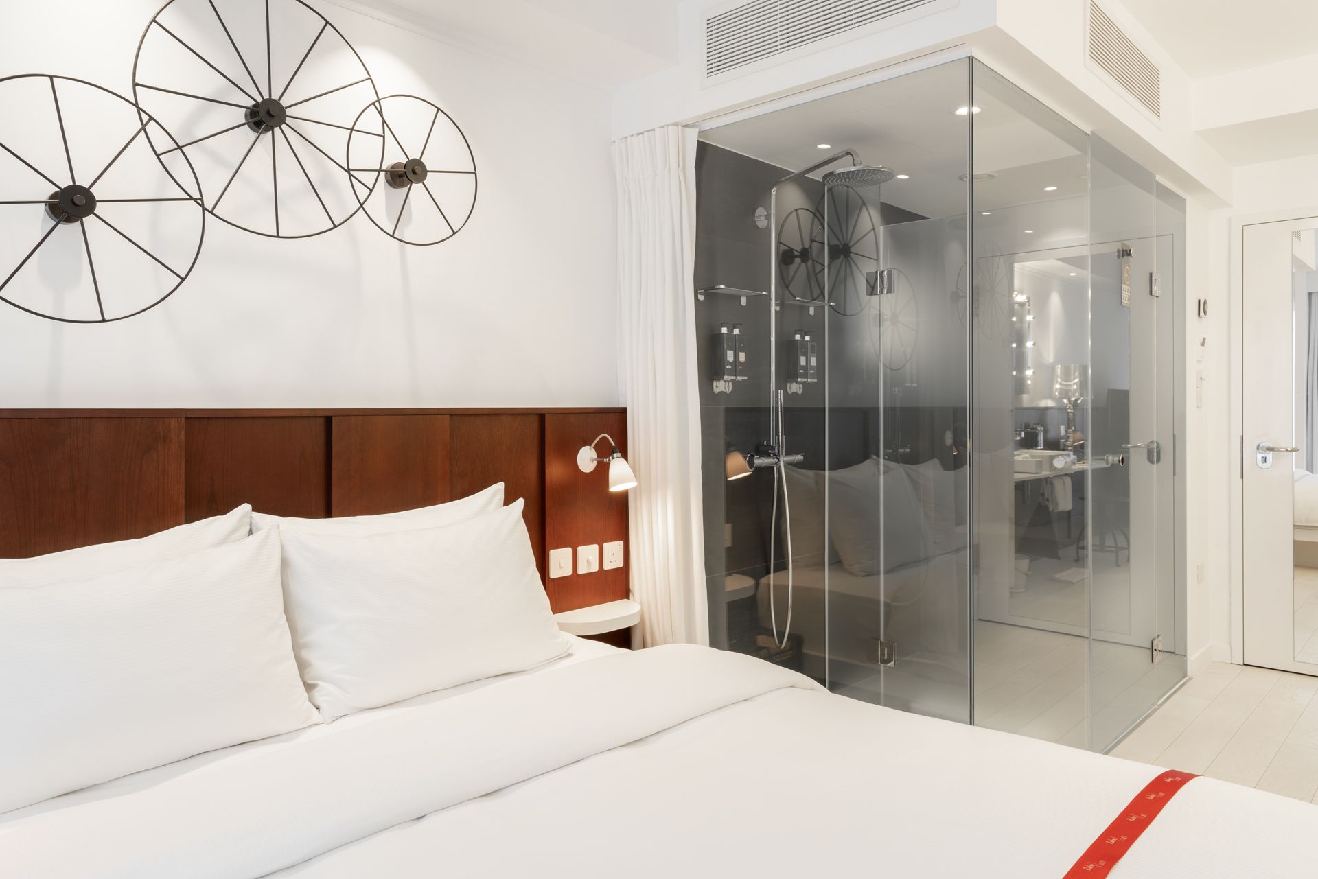 A modern hotel room with a large bed, a wooden headboard, a glass-enclosed shower, and a wall-mounted light fixture.