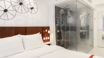 Camera d'hotel moderna con un letto grande, una testiera in legno, una doccia vetrata e un corpo illuminante fissato alla parete.