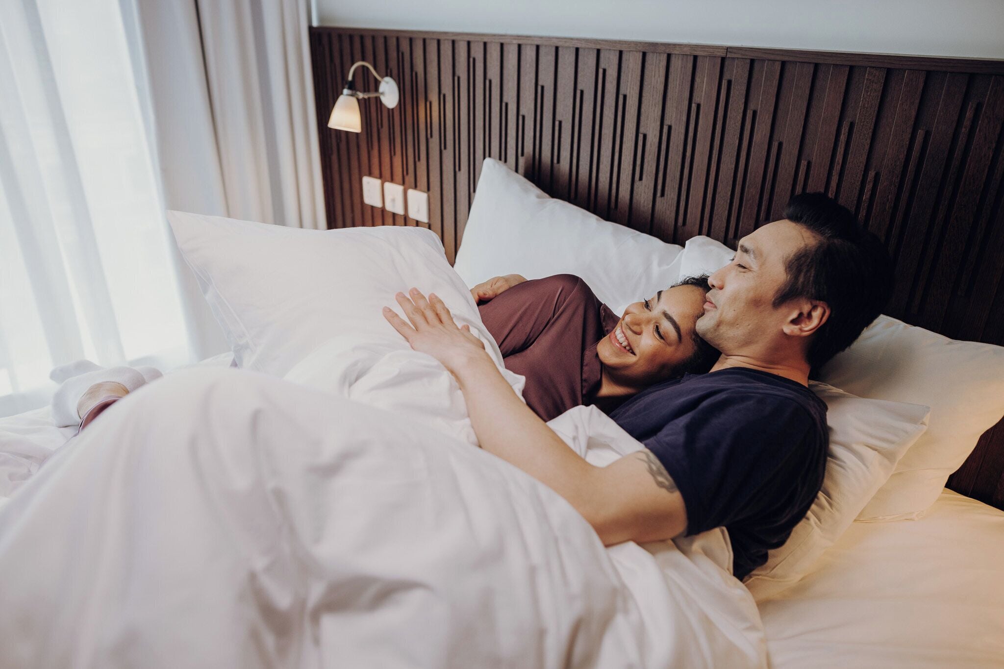 A couple lying in bed with a wooden headboard, white bedding, and a bedside lamp.
