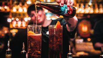 Eine Bar-Szene mit zwei hohen Gläsern, gefüllt mit einem dunklen Getränk, eines mit einer Orangenscheibe verziert. Eine Hand gießt aus einer mit 'Karma Cola' beschrifteten Flasche.